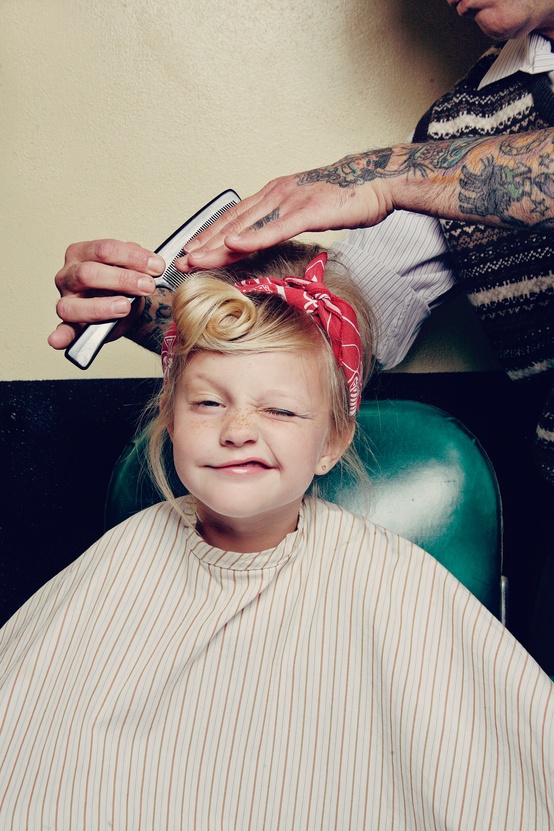 Cute little girl making funny face while getting a haircut by tattooed barbour